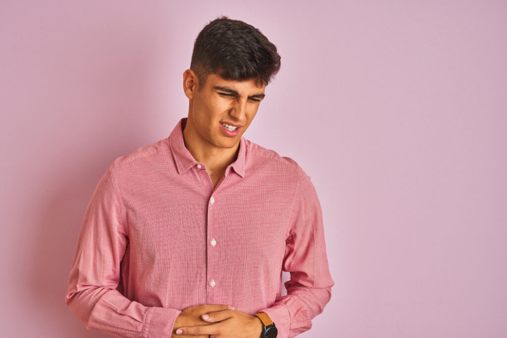 Young indian man wearing elegant shirt standing over isolated pink background with hand on stomach because indigestion, painful illness feeling unwell. Ache concept.
