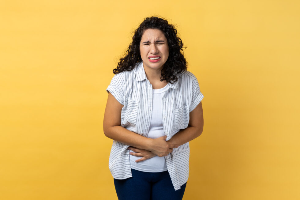 Portrait of sad depressed unhealthy woman with dark wavy hair suffering from strong stomachache, frowning, pain during period. Indoor studio shot isolated on yellow background.